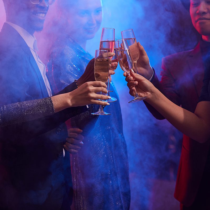 Cropped image of young people clinking champagne glasses in smoky night club while celebrating holidays at party, copy space
